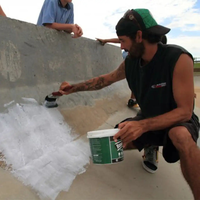 Person working on a concrete surface with a tool and bucket, another person observing in the background.