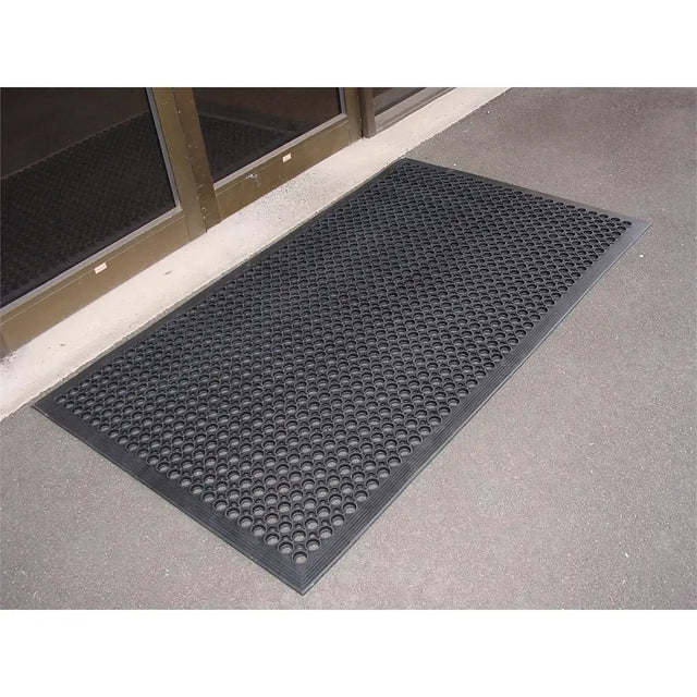 Black doormat with textured pattern on a concrete floor outside a building entrance.