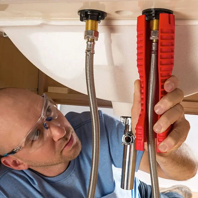 A plumber wearing safety glasses and a blue shirt works underneath a white sink, using a red-handled pipe wrench to adjust a metal water supply line near a black drain cap.