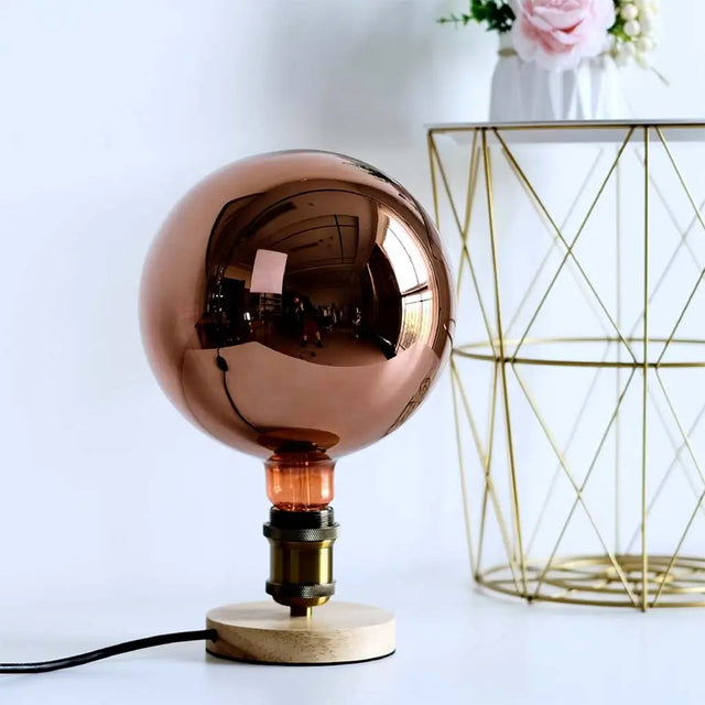 Copper spherical lamp on a wooden base with a geometric gold cage in the background.