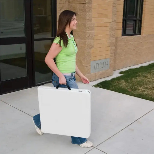 Woman in green shirt and blue jeans carrying a white suitcase outside a building.