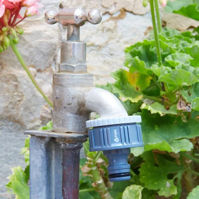 Metal garden faucet with grey connector attached, surrounded by green plants and flowers.