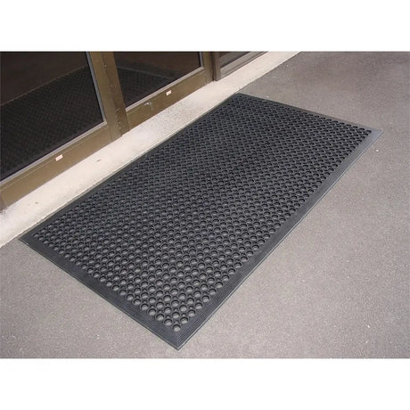 Black doormat with textured pattern on a concrete floor outside a building entrance.