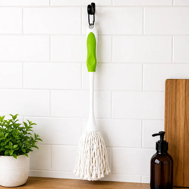 dish Mop with green handle hanging on a white tiled wall next to a plant and bottle.