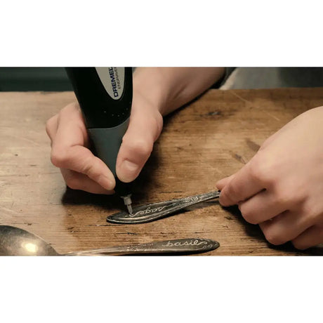 Person using a Dremel tool on a spoon with 'basil' engraved on it, on a wooden surface.