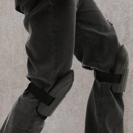 Close-up of a person wearing dark gray pants with black knee pads attached near both knees, shown against a light textured wall.