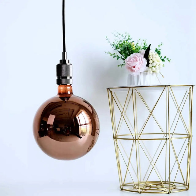 Copper pendant light bulb hanging above a gold geometric vase with flowers on a white background