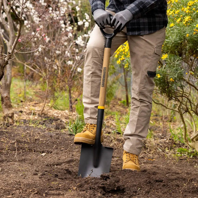 Person holding a spade in a garden with trees and flowers in the background