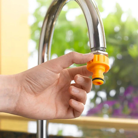 Hand holding an orange water filter nozzle attached to a silver faucet with a blurred green outdoor background.