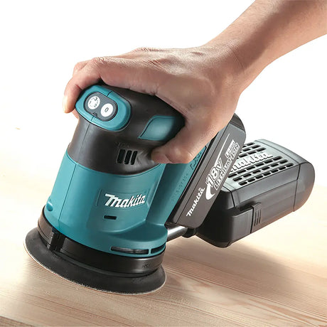 A Makita cordless orbital sander being used by a hand on a light wood surface, showing the teal and black tool with its battery attached.