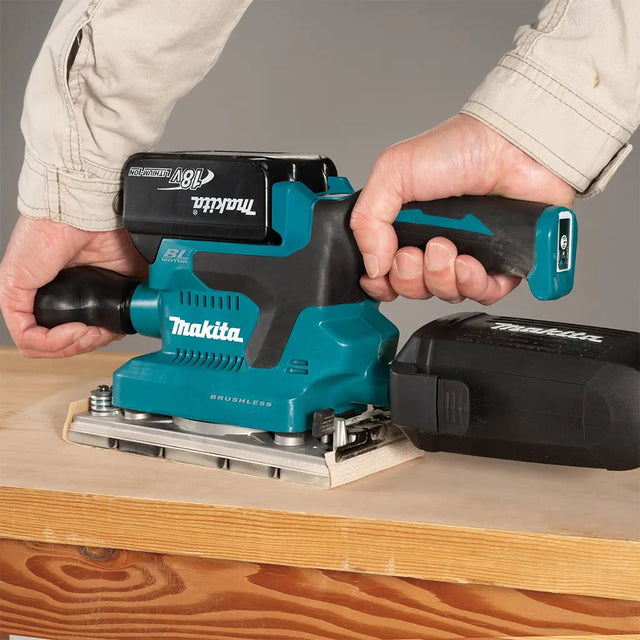 A person using a Makita cordless plunge router to sand a wooden board, with the teal and black router and a black battery pack visible.
