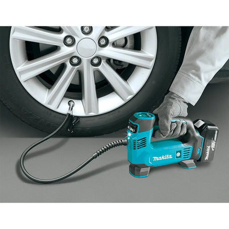 A Makita cordless tire inflator being used to inflate a car tire, with a person's gloved hand holding the handle and a battery attached. The Makita brand name is clearly visible on the tool.