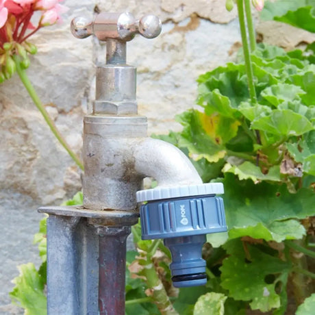 Metal garden faucet with grey connector attached, surrounded by green plants and flowers.