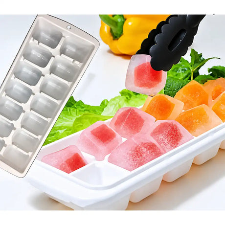 A white silicone ice cube tray shown with several translucent pink and orange ice cubes inside, plus a black pair of tongs lifting one cube. A second empty tray is visible at left, with lettuce, mint, and a yellow pepper in the background as styled props.