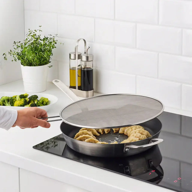 Person cooking food in a frying pan on a stove with a white tiled wall in the background.